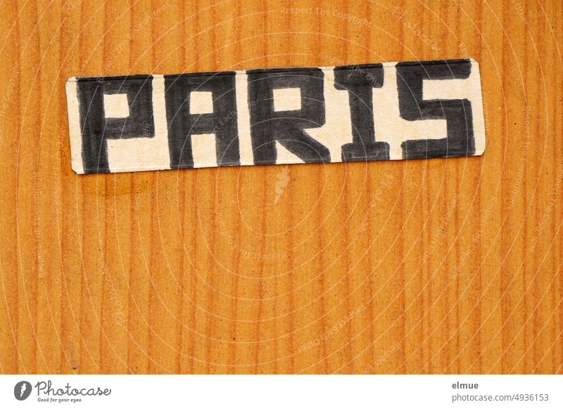 a cardboard sign with the inscription PARIS in black handwritten block letters is glued to a light brown wood texture wall Paris Piece of paper Handwriting
