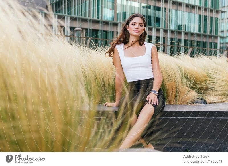 Gorgeous young female sitting on bench in modern city park woman self assured building portrait confident appearance street gorgeous casual long hair dark hair