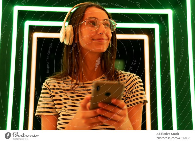 Young meloman using smartphone in neon corridor woman smile texting green orange listen music female young happy gadget device wireless browsing positive