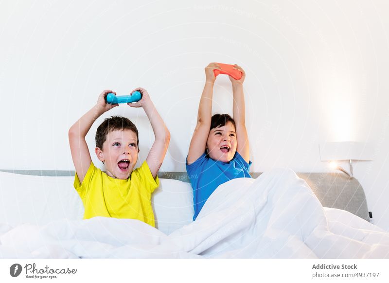 Children playing video game in bed children sibling gamepad arms raised videogame entertain cheerful amusement pastime leisure smile hobby sister brother boy