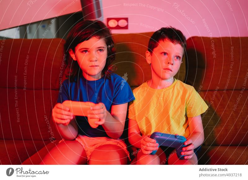 Children playing video game in evening time children sibling living room videogame entertain amusement pastime leisure couch sofa dim neon late barefoot hobby