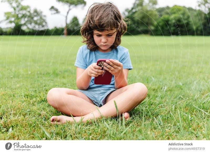Girl using smartphone on park lawn girl watch video unhappy upset weekend summer pastime child gadget sad device cellphone grass internet childhood angry online