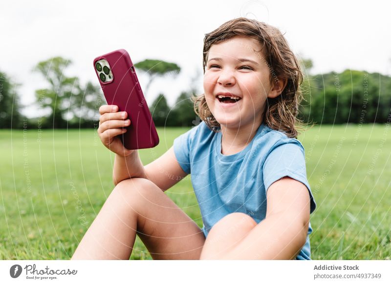 Cheerful girl using smartphone on park lawn watch video cheerful excited summer pastime happy child gadget device cellphone delight grass internet glad weekend