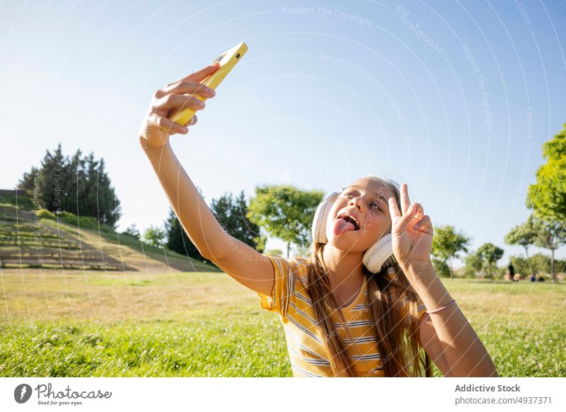 Joyful girl in headphones taking selfie in park teenage smartphone v sign photography social media music listen peace two fingers gesture tongue out show tongue