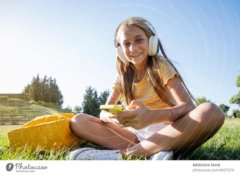 Smiling girl in headphones browsing smartphone in park teenage online music listen meloman pastime leisure lawn positive summer cellphone playlist gadget device