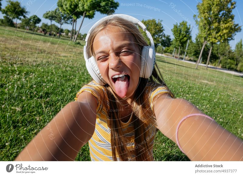 Joyful girl in headphones taking selfie in park teenage smartphone photography social media music listen gesture tongue out show tongue content carefree grimace
