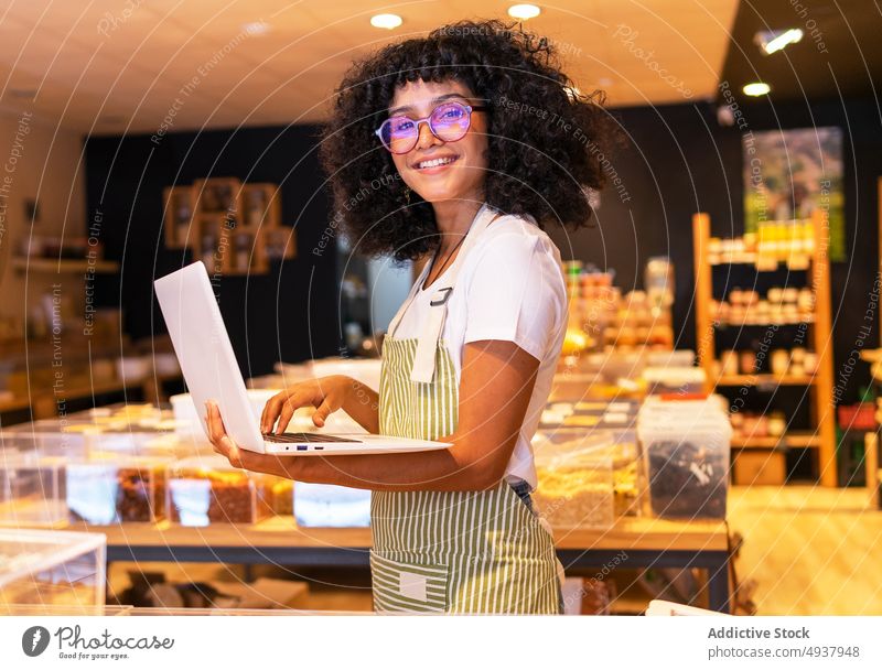 Cheerful black female vendor inventorying goods woman laptop using check store zero waste grocery young african american ethnic shop glass box small business