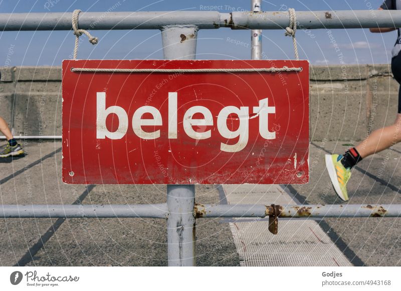 Sign with the inscription "Occupied" on a railing. In the background the feet of two joggers Signs and labeling Coated Jogging Mole Exterior shot Baltic Sea Sky