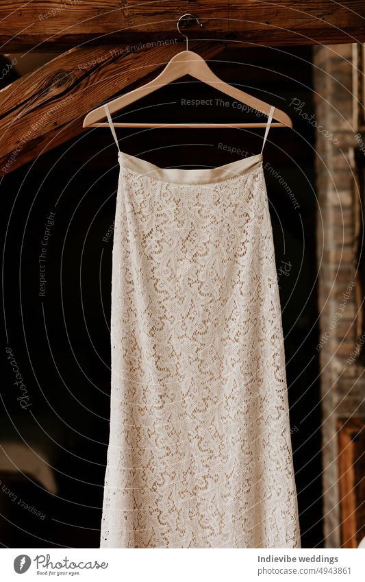 A beautiful lace wedding dress is hanging on a wooden hanger in the attic. Stunning white details, pretty style. Black background copy space. Big day preparation design.