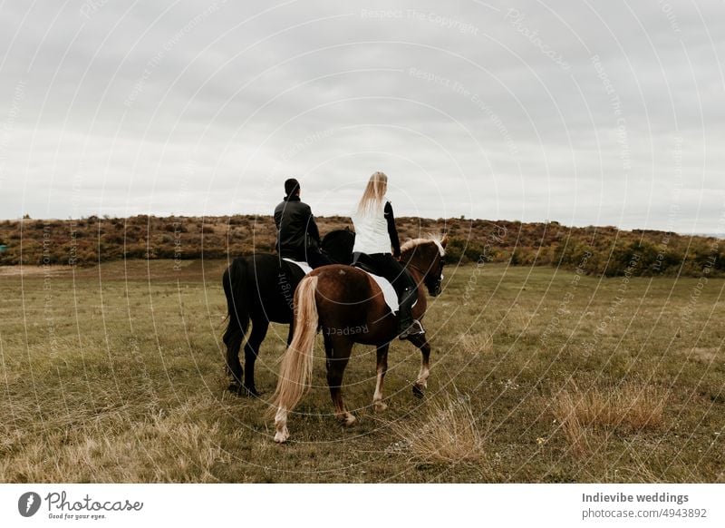 A couple is horse riding on a meadow facing the distance. Romantic and active outdoor date. Autumn landscape, with young couple enjoying a horse riding. Overcast weather, beautiful horses.