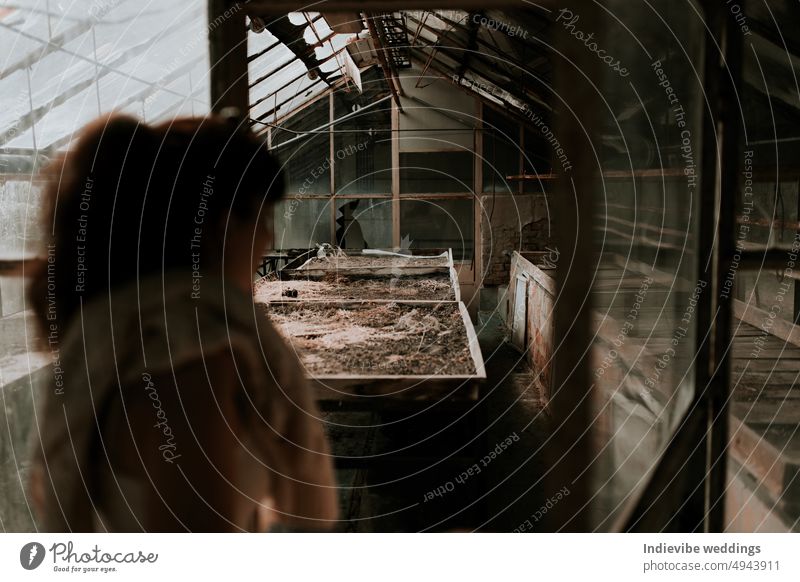 An abandoned greenhouse detail. A woman in the blurry foreground is entering the greenhouse. Rusty and dusty interior, broken glass. Old unused space, post apocaliptic scene. Copy space.
