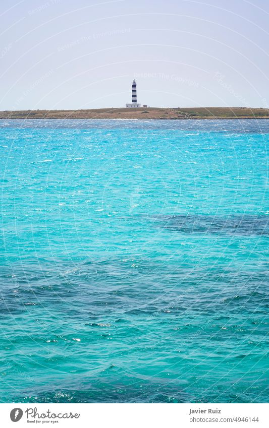 A turquoise sea in the foreground with some flat waves, in the background a blue lighthouse and blacco, vertical, space to copy island water balearic islands