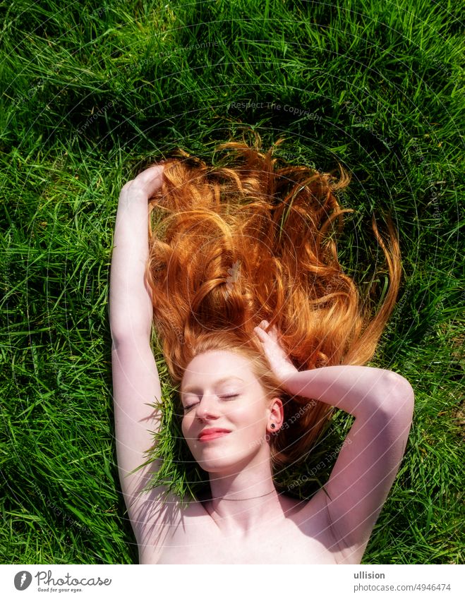 Portrait of a beautiful young sexy red-haired woman, lying in the spring sun in happiness, relaxing on the green grass, the red hair draped freely around the head.