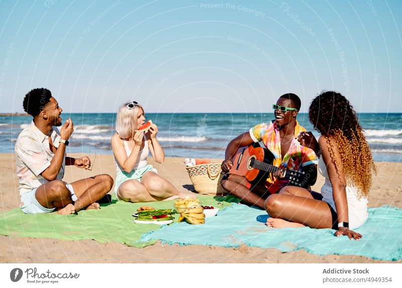 Diverse friends eating fruits and listening to guitarist play beach smile sea summer men women weekend picnic happy coast melody holiday music song together