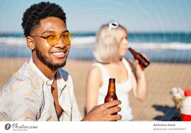 Black man drinking beer with friends beach sea picnic bottle together meeting weekend woman summer alcohol booze beverage diverse multiracial multiethnic smile