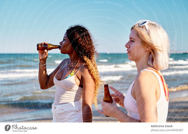 Young diverse ladies standing on seashore and drinking beer on summer day women beach friend holiday spend time admire together ocean young multiracial