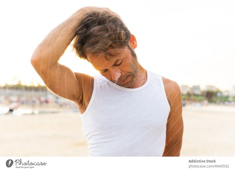 Middle aged man stretching neck on beach warm up training early summer bend exercise male mature middle age touch head morning workout wellbeing