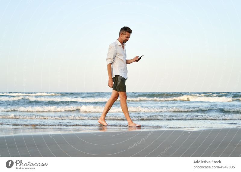 Businessman walking near sea and using smartphone businessman texting vacation beach summer telework male resort barefoot coast device gadget shore white shirt