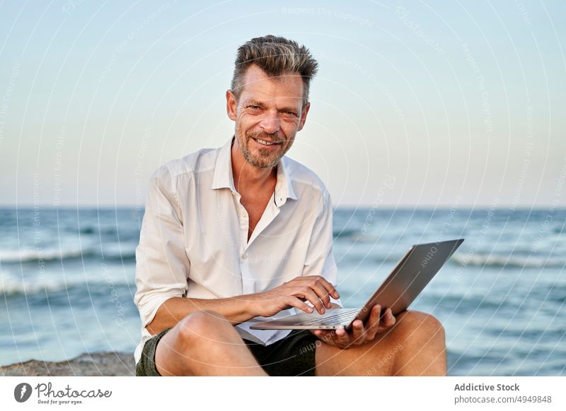 Mature businessman working on rocks near sea laptop using smile data telework male mature middle age independent project sunset happy smart casual positive