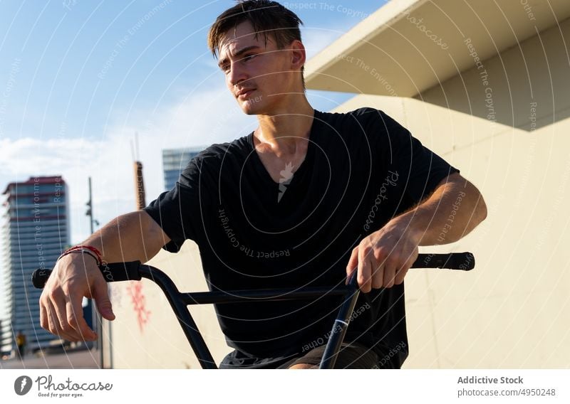Young man with bicycle in skate park wall hobby weekend break daytime modern urban male young casual t shirt summer bike handlebar touch pause building cyclist