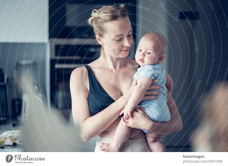 Pretty young mother holding her newborn baby boy standing near kitchen window at home. woman family childhood adult care motherhood mom room parent love white