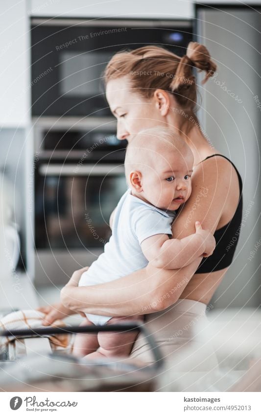 Woman wiping kitchen sink with a cloth after finishing washing the dishes while holding four months old baby boy in her hands. childhood family motherhood woman