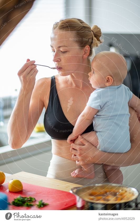 Woman cooking and tasing food while holding four months old baby boy in her hands. childhood family motherhood woman parent kitchen home son busy learning