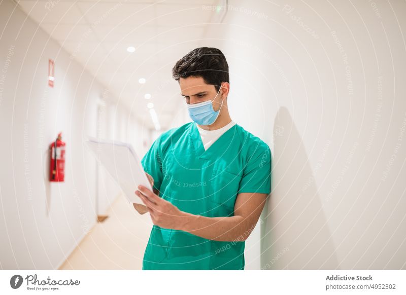 Attentive doctor with papers in hospital corridor read uniform professional job sterile attentive man surgeon light lamp shiny focus medic mask protective