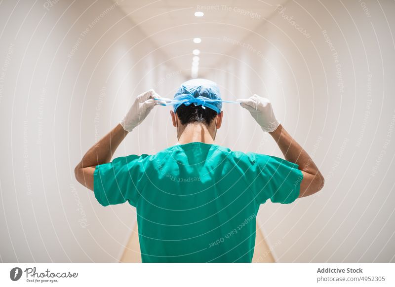 Anonymous surgeon tying medical cap in clinic corridor uniform prepare professional man hospital work protect sterile glove passage narrow latex material lamp
