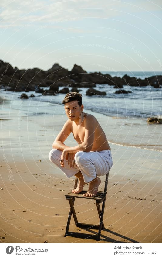 Shirtless man sitting on knees on chair near sea beach style stand balance concept vulnerable embrace male model young shirtless barefoot coast shore ocean
