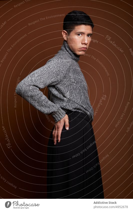 Stylish homosexual model in sweater on brown background gay fashion style feminine lgbt man unemotional portrait knitted cloth emotionless gaze rest stand