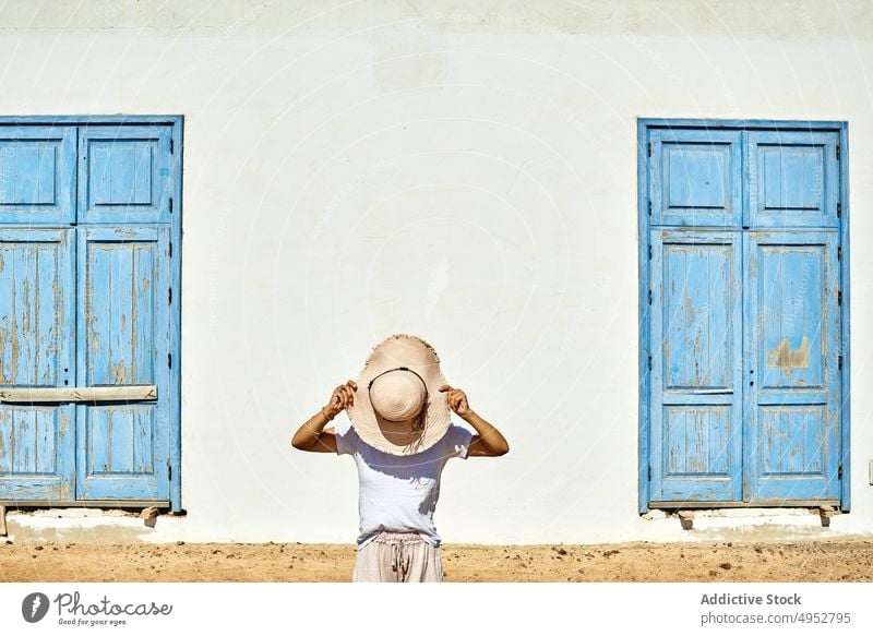 Anonymous woman covering face with hat against rural cottage cover face hide incognito house facade shutter exterior white blue architecture residential