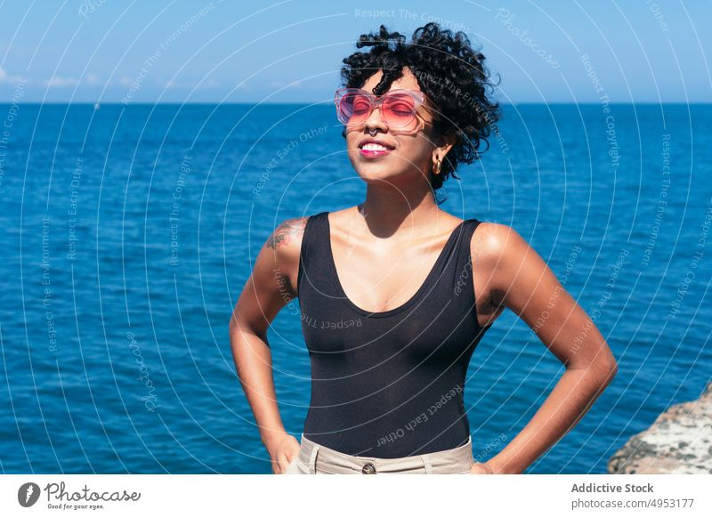 Smiling black woman in sunglasses against endless sea cheerful individuality style tattoo feminine hand on waist blue sky modern creative design