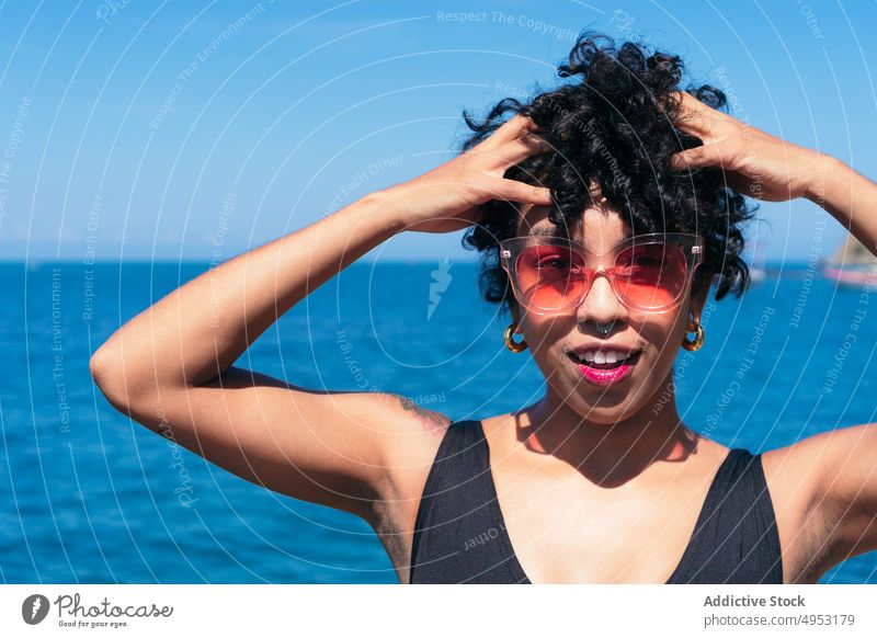 Smiling black woman in sunglasses against endless sea cheerful individuality style tattoo feminine hands on head blue sky modern creative design