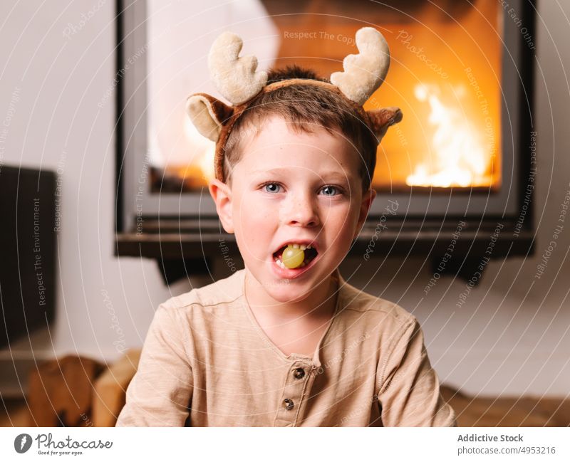 Cute boy in reindeer antlers eating grape kid headband christmas fireplace fruit living room holiday occasion xmas home festive celebrate childhood adorable