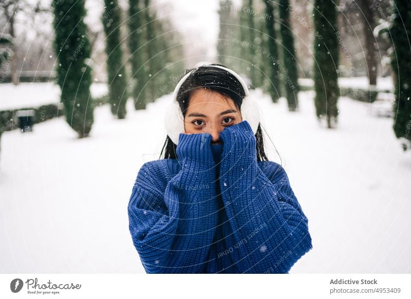 Millennial Asian woman in knitwear in winter park cover face shy snowfall gentle cold portrait weather wintertime idyllic urban tender timid style warm clothes
