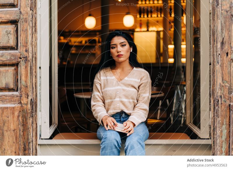 Asian woman in earbuds resting on cafe windowsill feminine style casual romantic dreamy wooden material old weathered contemplate wireless gentle charming