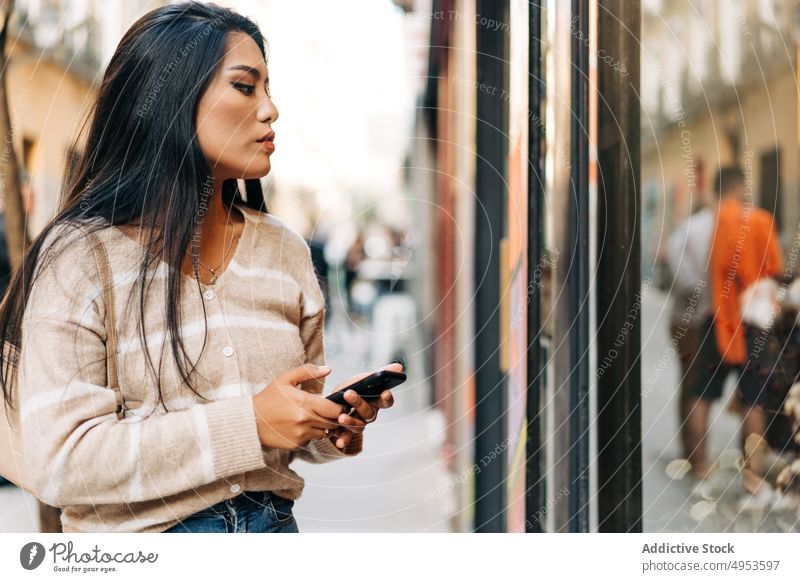 Asian woman with smartphone against glass wall in town reflection thoughtful feminine style casual pavement city attentive millennial charming focus cellphone