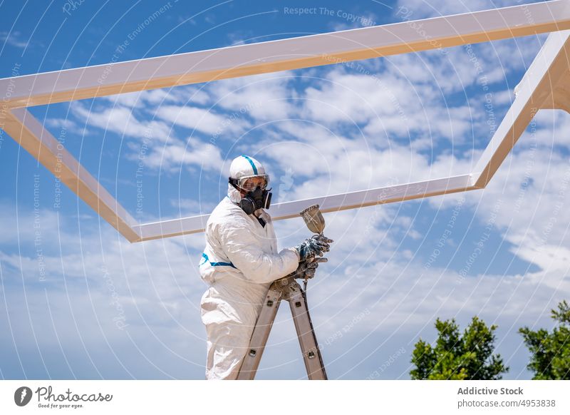 Unrecognizable worker painting building on sunny day air brush uniform protect mask sky cloudy daytime ladder job labor employee equipment structure frame