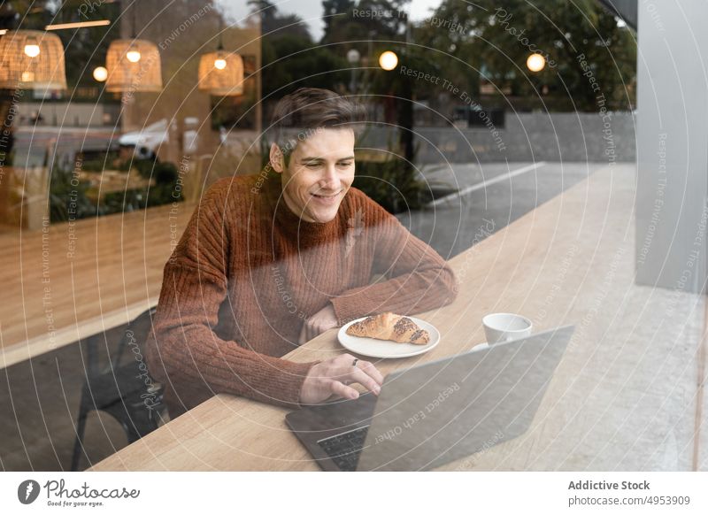 Cheerful man with laptop talking on smartphone in cafe coffee happy video call window young student freelance break breakfast cup croissant cafeteria device
