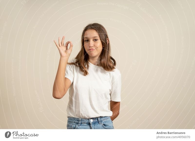 Portrait of cheerful brunette teenager in white t-shirt showing ok sing isolated over beige background. pretty portrait girl young female cute beautiful smiling