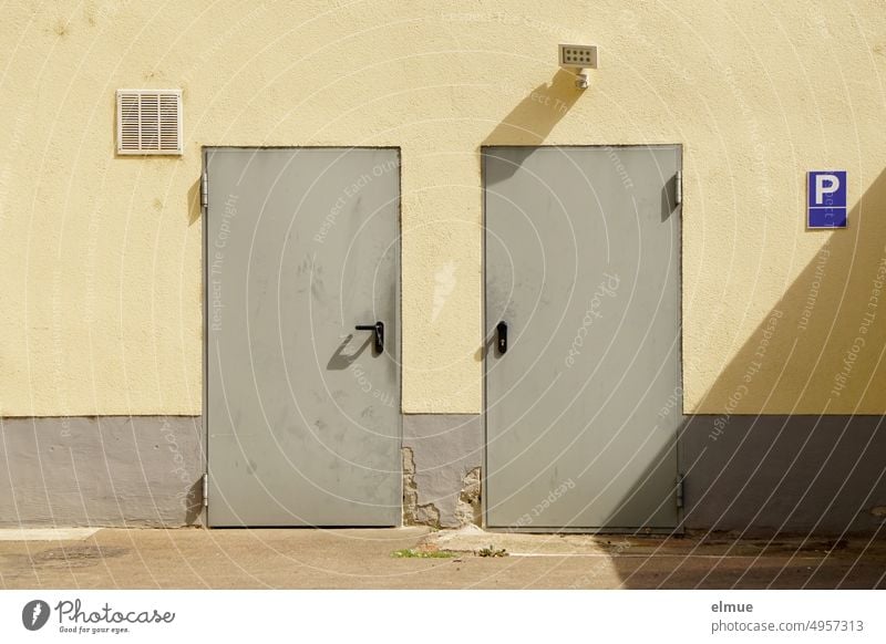 two gray, worn metal doors lead into a functional building on the wall of which there is a fan, a small spotlight and a blue parking sign Metal door Entrance