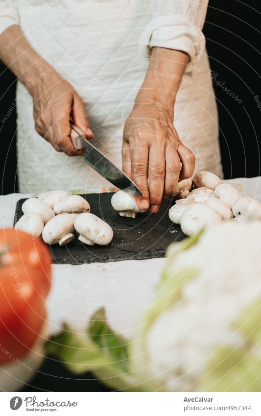 Wrinkled hands of a senior woman cutting mushroom on a black board with the sleeves rolled up.Teaching the correct way to chop,cooking a vegetarian meal also with tomato and cauliflower