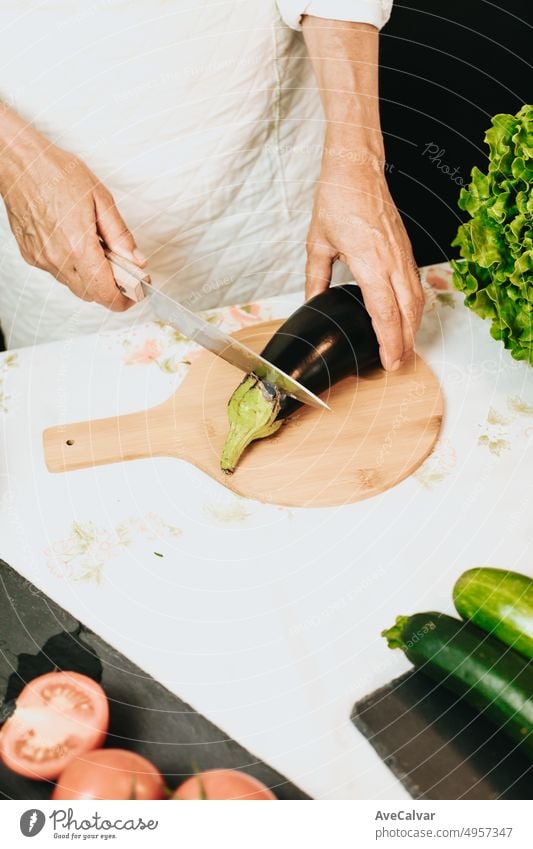 Older woman chef cooking a vegan recipe for a television show. Hands holding a knife and cutting an eggplant combined with zucchini,tomatoes and lettuce on a wood table.Balanced and healthy meal