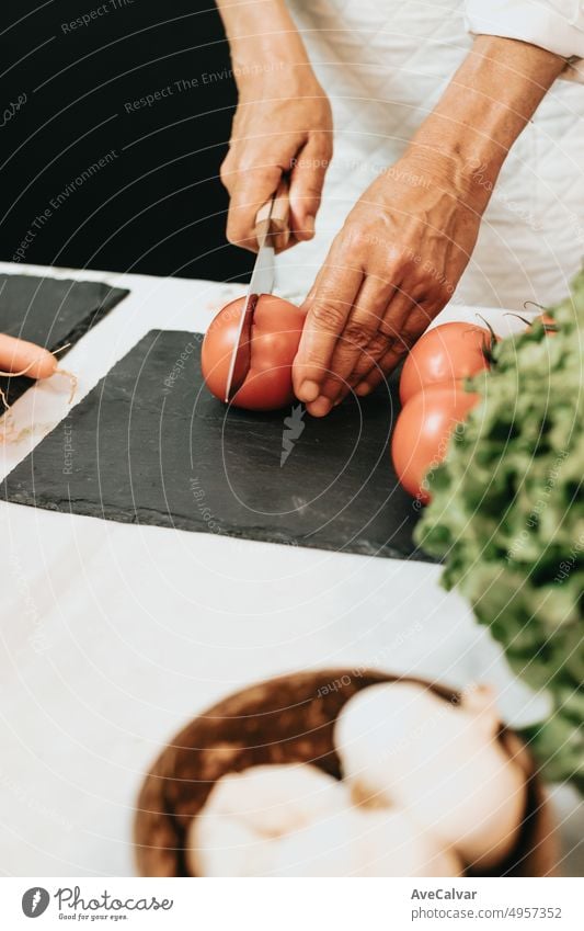 Old woman teaching a cooking recipe with a white kitchen apron. Close up of hands cutting tomatoes with a knife on a black board preparing a healthy,delicious and complete meal for lunch and dinner