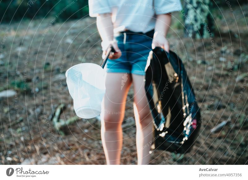 Young woman picking up trash with garbage tongs pointing at camera.Forest background.Plastic cups and garbage bag.New generation.Recycle, protecting the planet, environmental conservation,volunteer