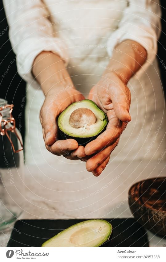Woman chef holding and showing ripe cut avocado on white kitchen apron.Half avocado on black board,wooden bowl for fresh guacamole and glass jar for healthy and nutritional avocado smoothie