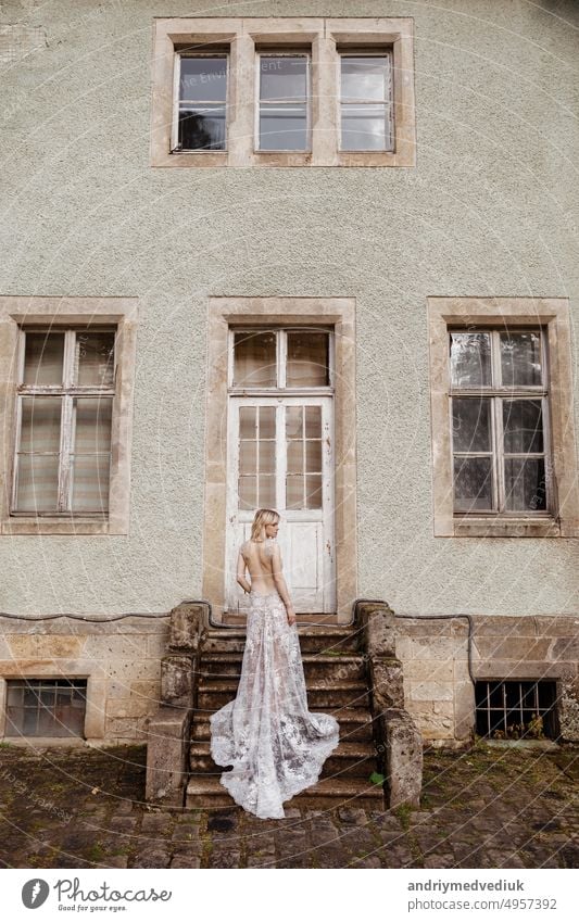 fashion outdoor photo of gorgeous young bride with blond hair in elegant long wedding dress posing near old castle. back view woman model style girl lady