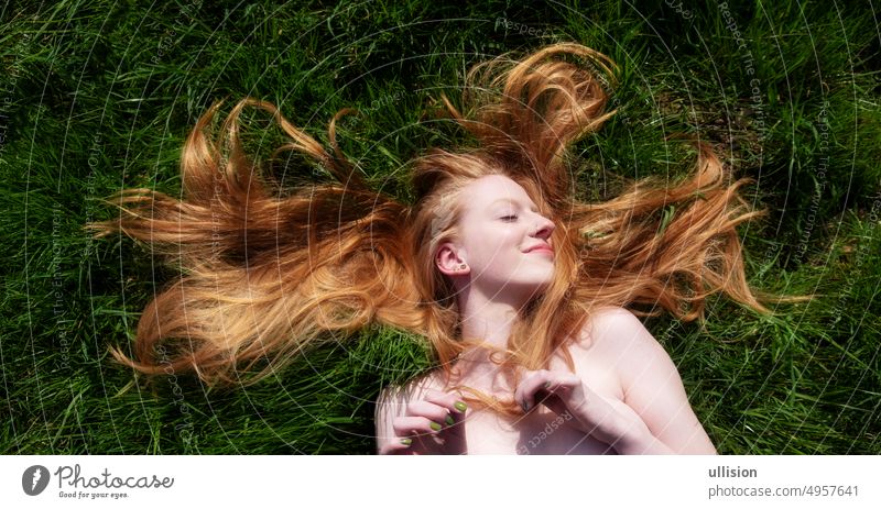 Portrait of a beautiful young sexy red-haired woman, lying in the summer in the sunshine in happiness, lying relaxed on the green field, grass, the red hair draped around the head.