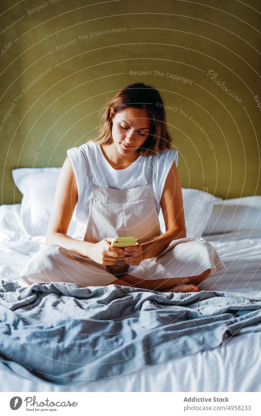 Young woman text messaging on smartphone while sitting on bed using text message social media cellphone connection surfing mobile female gadget domestic